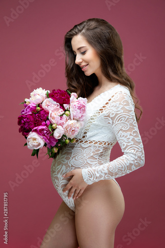 A beautiful pregnant woman with a bouquet of peony flowers and with long hair, dressed in lace white lacy body suit