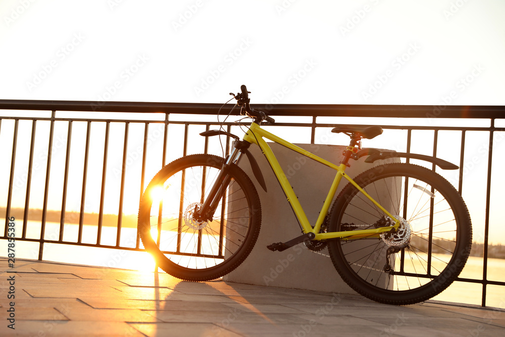 Yellow bicycle parked near railing on city waterfront at sunset - obrazy, fototapety, plakaty 