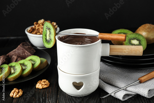 Fondue pot with chocolate and kiwis on black wooden table