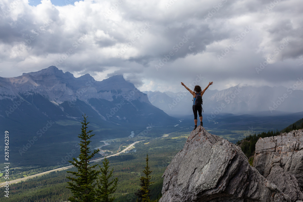 Adventurous Caucasian Girl with open arms is on top of rocky mountain during a cloudy and rainy day. Taken from Mt Lady MacDonald, Canmore, Alberta, Canada.