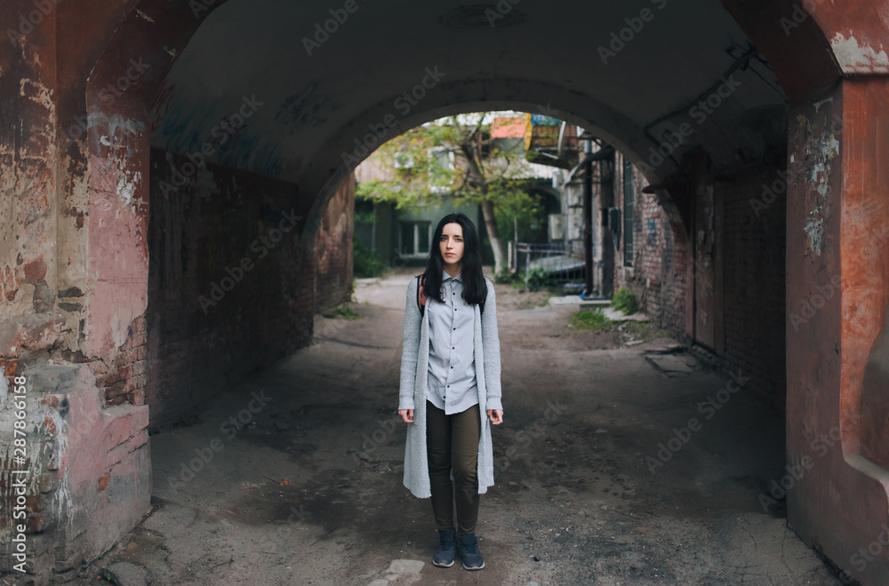 A young european sad girl in a gray cardigan and a shirt stands in the arch of an old slum yard and looks at the viewer. The concept of poverty, decline and lack of prospects.