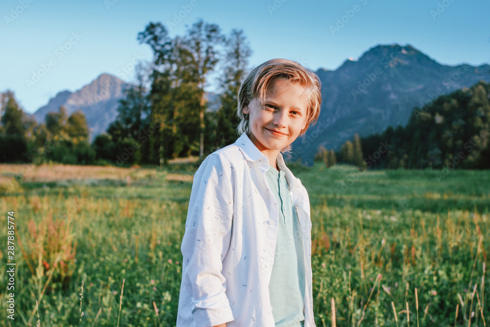 Blonde smiling boy looking at camera on background of wonderful view of the green meadow and mountains, family travel adventure lifestyle