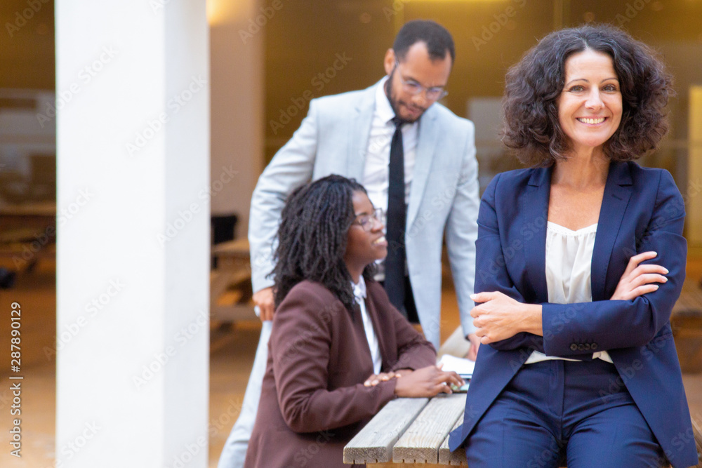 Fototapeta premium Happy confident business lady posing with her team in street cafe. Her colleagues sitting and standing at table and talking in background. Teamwork outside concept