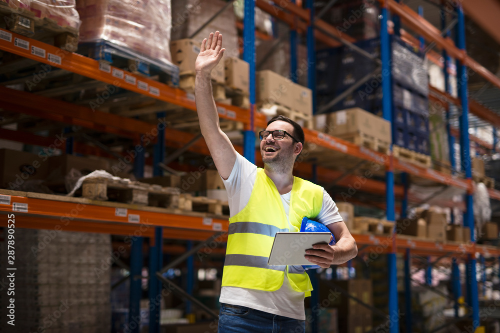 Warehouse worker with tablet and protective uniform standing between shelves in storage center and waving to his coworker. Warehouse worker with tablet and protective uniform standing between shelves in storage center and waving to his coworker.