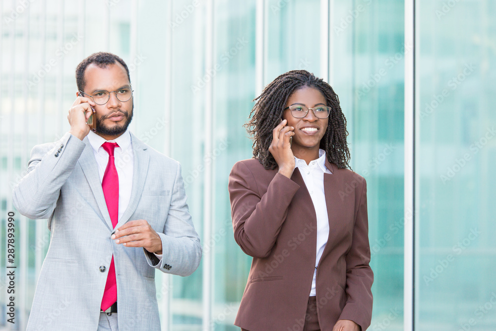 Excited business colleagues discussing project on mobile phone on their way to office. Business man and woman walking outside and talking on cellphones. Negotiation on phone concept Excited business colleagues discussing project on mobile phone on their way to office. Business man and woman walking outside and talking on cellphones. Negotiation on phone concept