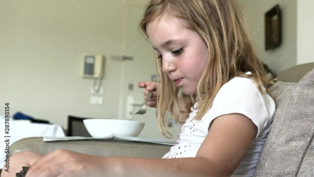 Little cute blonde girl sitting on a beige leather sofa at home and plays with digital tablet and eats pasta at the same time. 4K