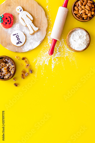 Cooking halloween cookies in shape of spooky figures, rollin pin, nuts and flour on yellow background top view copyspace