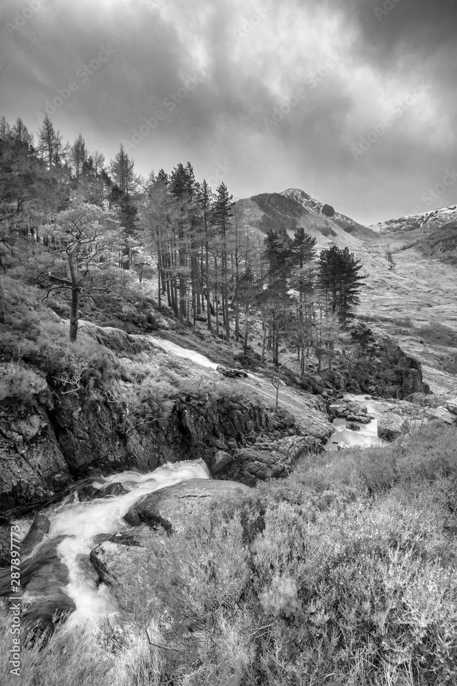 Stunning landscape image of Ogwen Valley river and waterfalls during Winter with snowcapped mountains in background in black and white
