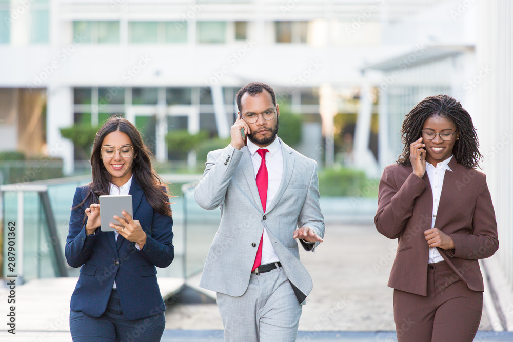 Multiethnic business team with gadgets going along urban glass facade. Business man and women walking outside, using tablet, talking on phone. Teamwork and communication concept