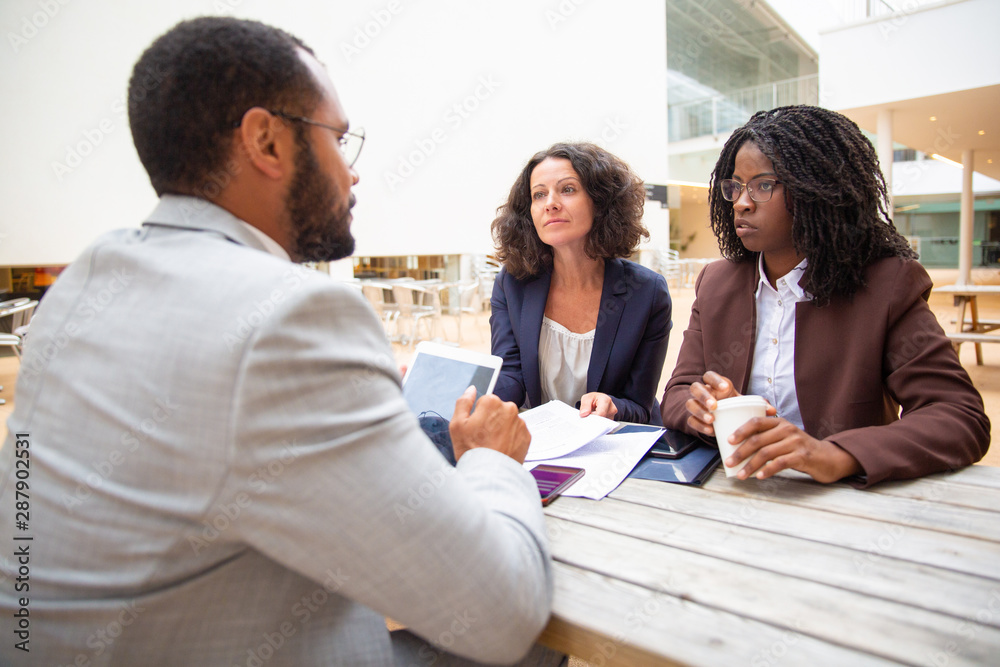 Fototapeta premium Diverse business women discussing deal with partner in cafe. Business man and women sitting at table, reading documents, drinking coffee, using tablet. Deal concept