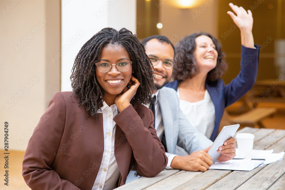 Business team of three relaxing in street cafe. Business man and women sitting at table outside, waving hello, looking at camera and smiling. Successful business team concept Business team of three relaxing in street cafe. Business man and women sitting at table outside, waving hello, looking at camera and smiling. Successful business team concept