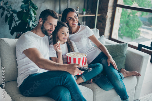 Portrait of nice attractive lovely charming cheerful cheery family wearing casual white t-shirts jeans denim sitting on divan having fun watching series serial video switching channel indoors