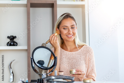 Beauty woman applying makeup. Beautiful girl looking in the mirror and applying cosmetic with a big brush. Girl gets blush on the cheekbones. Powder, rouge