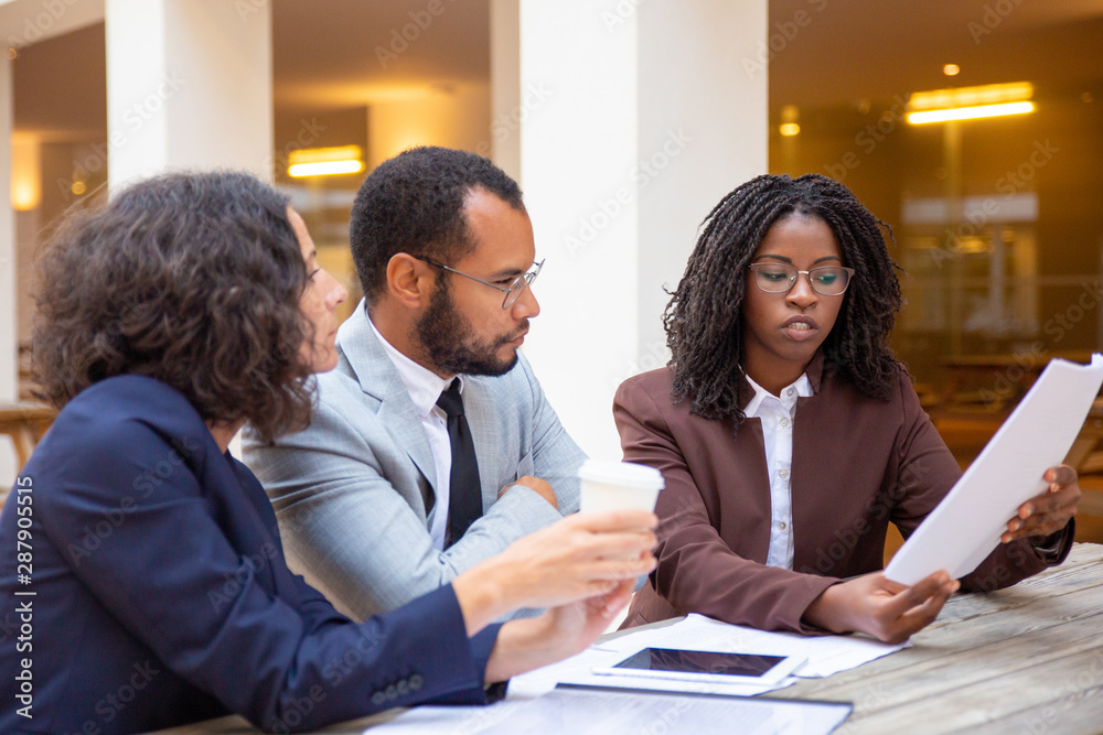 Legal expert explaining contract specifics to two business people in street cafe. Business man and women sitting at table outside, drinking coffee, reviewing papers and talking. Expertise concept Legal expert explaining contract specifics to two business people in street cafe. Business man and women sitting at table outside, drinking coffee, reviewing papers and talking. Expertise concept