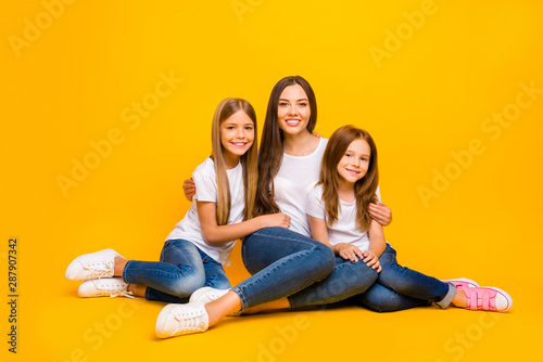 Two small ladies hugging young and beautiful babysitter sitting floor wear casual white t-shirts isolated yellow background