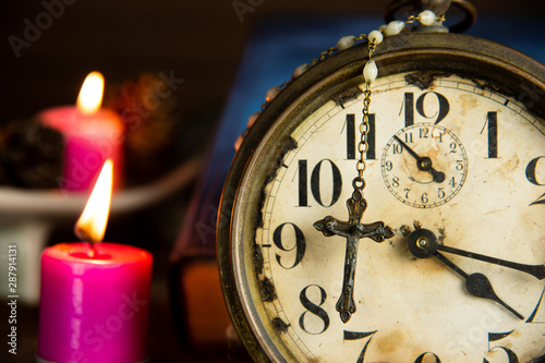 Beautiful antique Jesus crucifix and rosary beads hanging on vintage alarm clock with holy bible and light candles background.