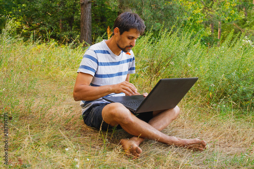 A young man sits on the grass with a flower in his mouth and works correspondence on a laptop.