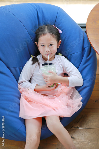 Portrait of happy little Asian child girl drinking iced Matcha Green Tea lying on sofa at cafe. photo