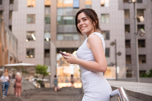Beautiful brunette business woman in grey smark casual dress working on a mobile phone in her hands outdoors. European city on background. copy space © AnnaDemy