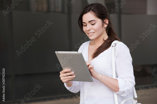 Beautiful brunette business woman in white skirt and grey suit trousers working on a tablet in her hands outdoors. European city on background. copy space
