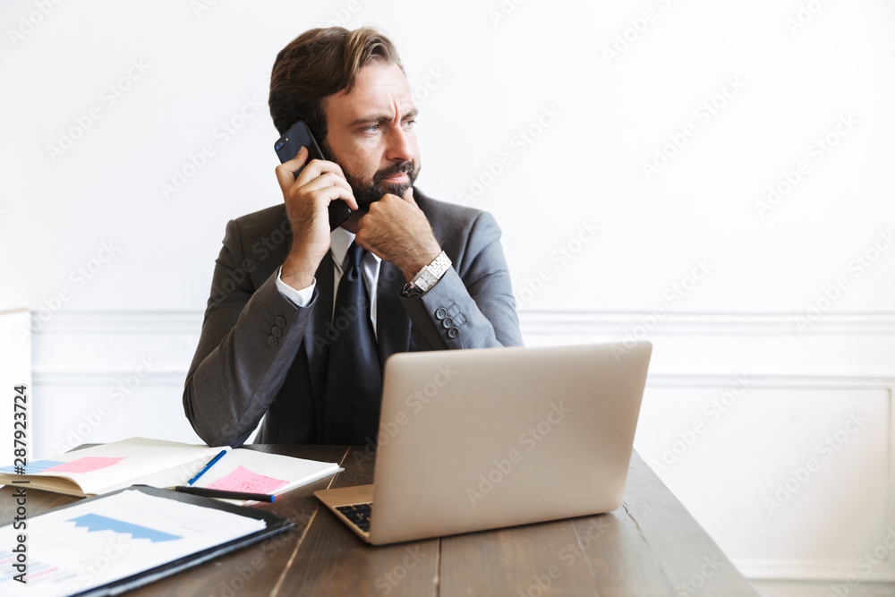 Serious thinking bearded business man sitting in office using laptop computer working indoors talking by mobile phone. Serious thinking bearded business man sitting in office using laptop computer working indoors talking by mobile phone.