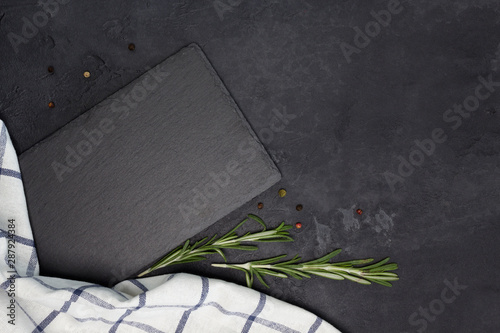 Slate plate, kitchen towel, rosemary, pepper on black concrete background. Cooking, recipe concept. Top view, flat lay, mock up, copy space