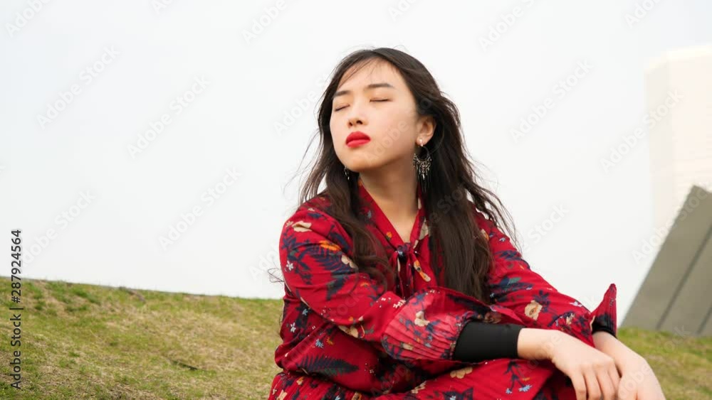 Portrait of a beautiful Chinese young woman in red dress sitting on grass field with eyes closed, enjoy the breeze blowing her black long hair, 4k footage, slow motion. Portrait of a beautiful Chinese young woman in red dress sitting on grass field with eyes closed, enjoy the breeze blowing her black long hair, 4k footage, slow motion.