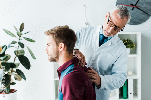 bearded chiropractor in white coat and glasses touching back of handsome patient in clinic