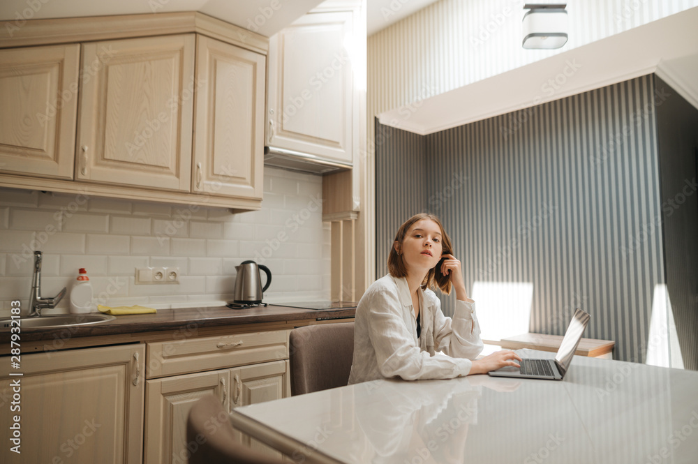 Portrait of a cute girl wearing a white shirt, sitting at a table in the kitchen, using a laptop and looking at the camera. Girl freelancer works at home in apartment on laptop. Portrait of a cute girl wearing a white shirt, sitting at a table in the kitchen, using a laptop and looking at the camera. Girl freelancer works at home in apartment on laptop.
