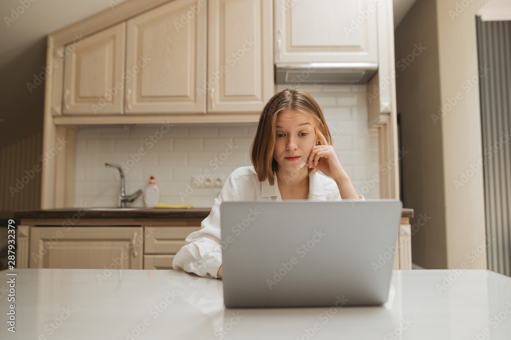 Portrait of surprised girl sitting at home in the kitchen at table, looking at laptop screen with shocked face. Surprised girl working on laptop at home in kitchen in apartment, wearing white shirt Portrait of surprised girl sitting at home in the kitchen at table, looking at laptop screen with shocked face. Surprised girl working on laptop at home in kitchen in apartment, wearing white shirt