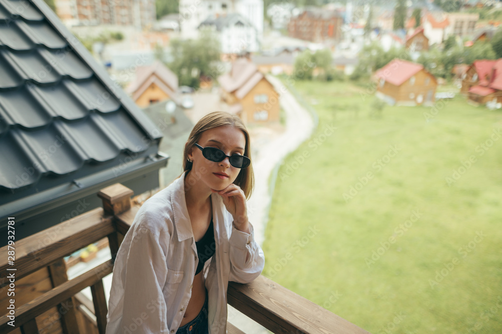 Stylish girl posing on the balcony in the hotel room, wears sunglasses, looks into the camera with a serious face. Beautiful woman standing on balcony in country house in mountains.