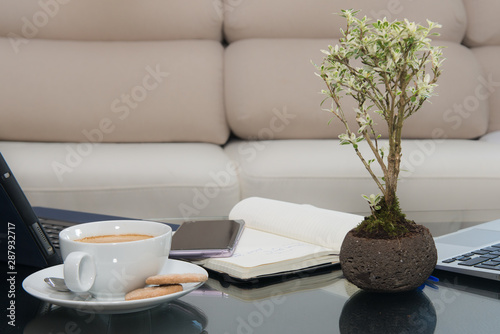 Glass table with a tablet, a cell phones notebook, a cup of coffee and a plant and in the background an elegant cream armchair