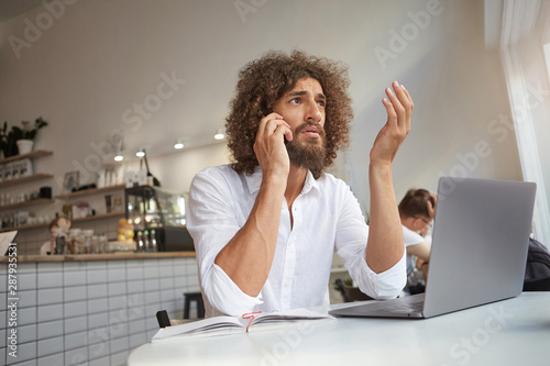 Indoor portrait of beautiful young freelancer with beard working remotely in public place, gesturing with hand while having serious conversation on phone