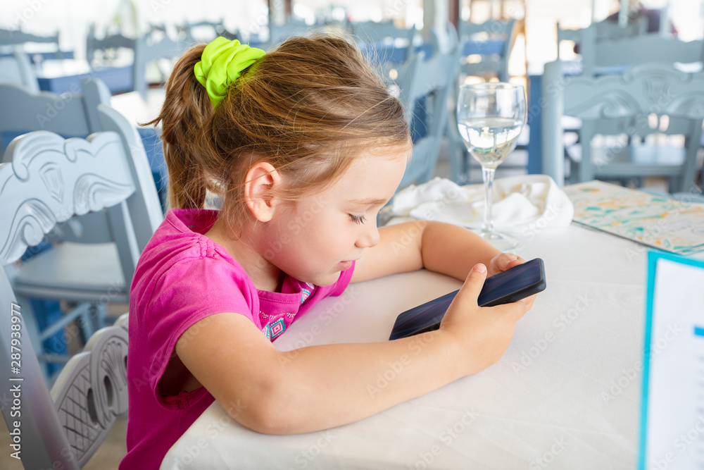 four years old blonde caucasian girl in summer, pink shirt, sitting in a restaurant, smiling and watching smartphone or mobile phone