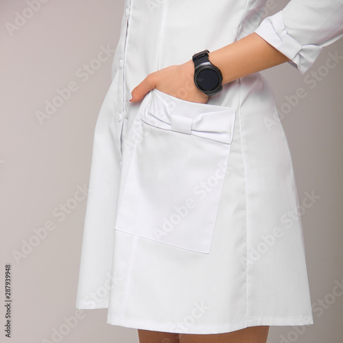 close-up young stylish nurse with one hand with wrist watch in the pocket of her medical dress on white wall background. medical fashion concept. free space