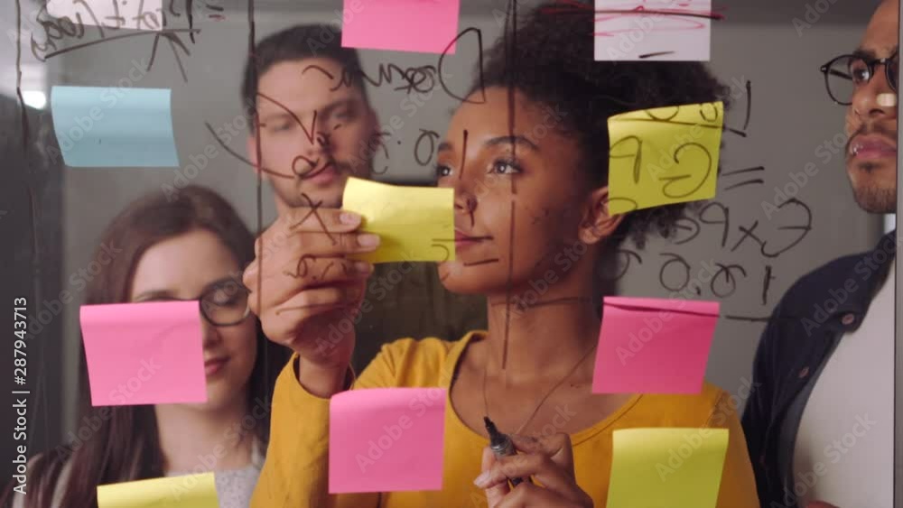Professional female executive standing with her colleague writing down business ideas on sticky notes and glass board at workplace