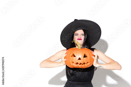 Young brunette woman in black hat and costume on white background. Attractive caucasian female model. Halloween, black friday, cyber monday, sales, autumn concept. Holding pumpkin. Look mystic.