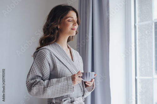 Portrait of pretty woman in bathrobe enjoying morning standing by the window and holding cup of tea. photo