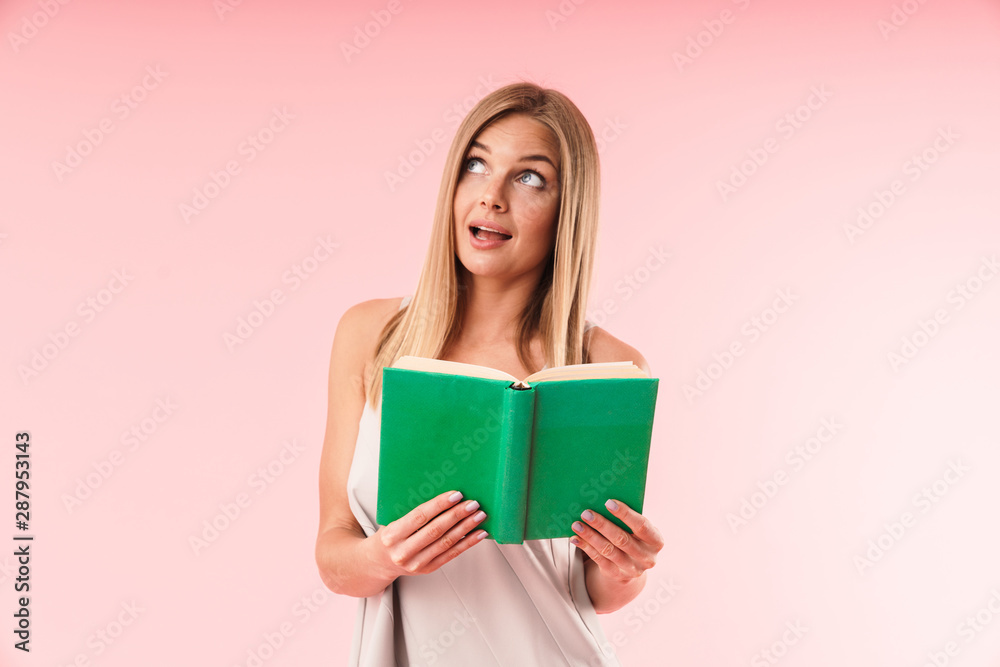 Image of joyful blond woman wearing dress smiling and looking upward while reading book Image of joyful blond woman wearing dress smiling and looking upward while reading book