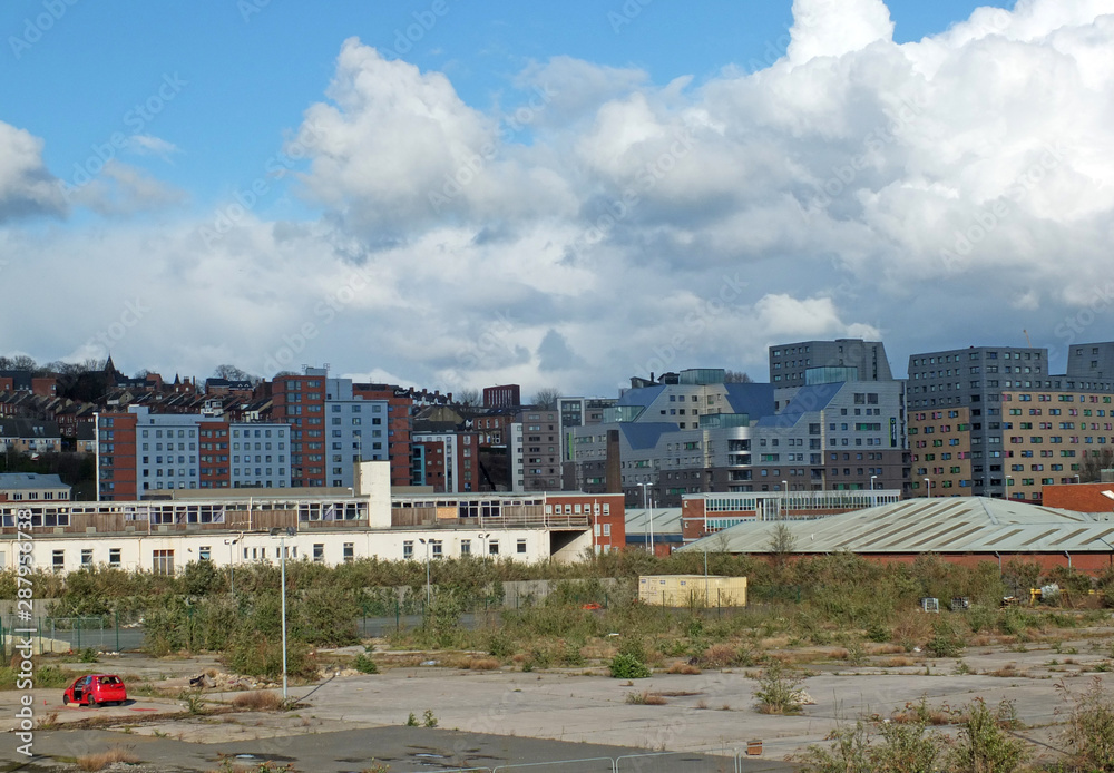 a large empty brownfield site in leeds england with weeds growing through concrete and a burned out abandoned car surrounded by city apartment buildings