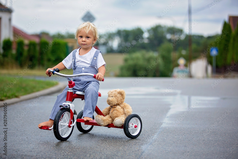 Cute little boy, with teddy bear toy, riding tricycle on the street in the rain, barefeet Cute little boy, with teddy bear toy, riding tricycle on the street in the rain, barefeet