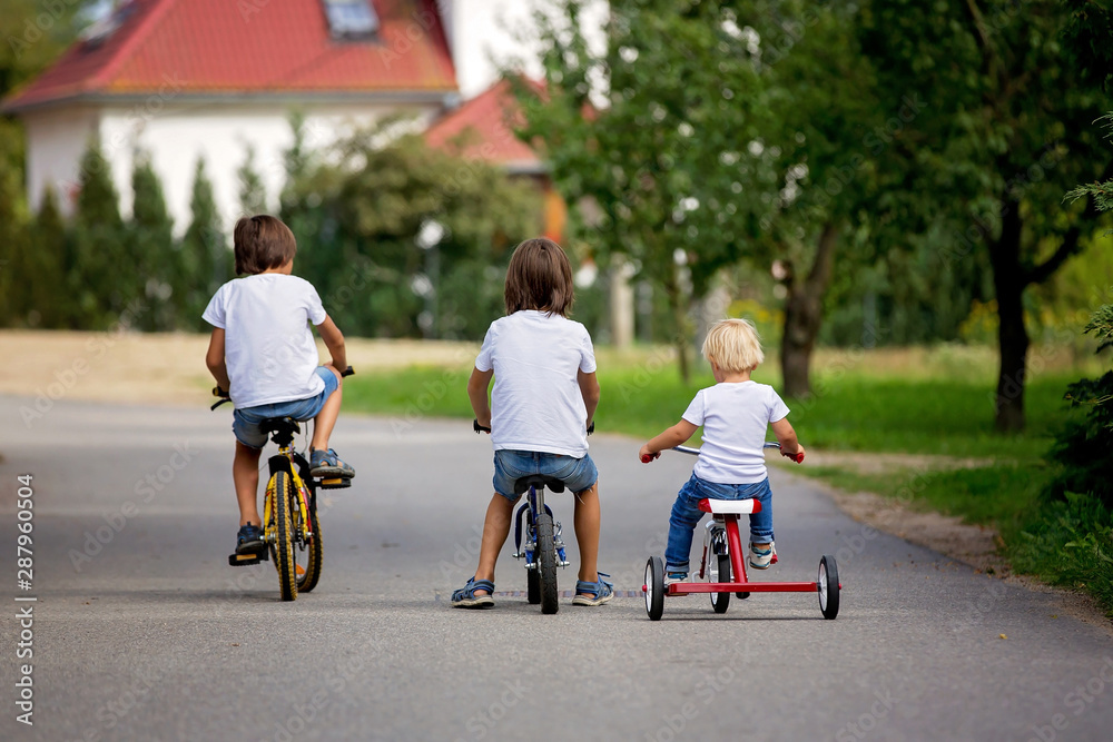 Three children, riding bike, tricycle and balance bike on the street in quiet village Three children, riding bike, tricycle and balance bike on the street in quiet village