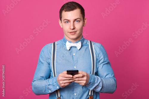 Horizontal shot of handsome young guy holds his mobile telephone, looks at camera with calm facial expression, male wears shirt, suspenders, white bow tie, poses in studio isolated on pink background. © sementsova321