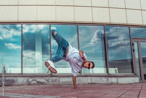 strong sporty man dancing break dance, stands one arm red-haired, young guy, motivation and lifestyle, summer city background of glass windows of building, jeans, sunglasses white T-shirt