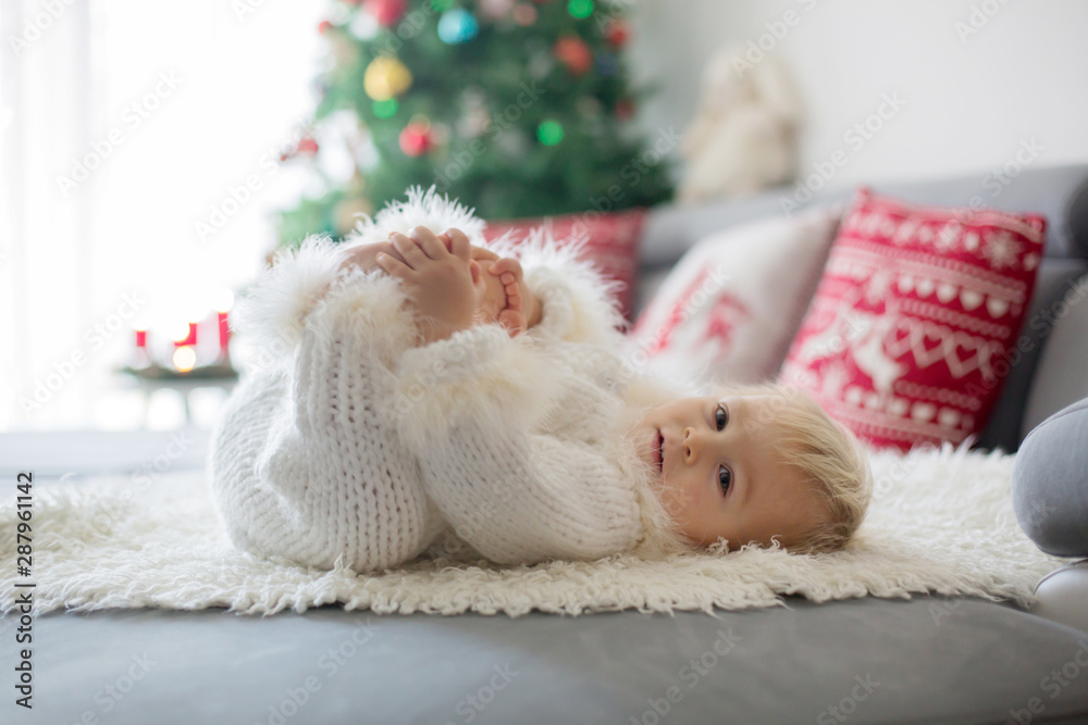 Curious little toddler boy, dressed in handknitted white overall, lying on the couch playing with gifts Curious little toddler boy, dressed in handknitted white overall, lying on the couch playing with gifts