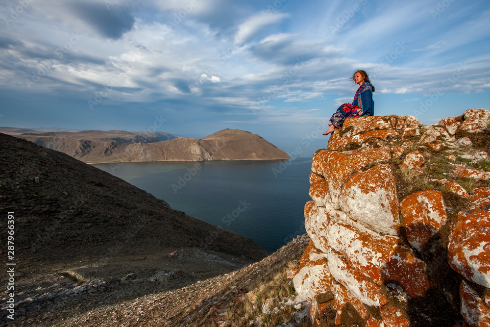 A girl sits on the stones of a high rock near Lake Baikal against the sky with beautiful clouds. European appearance. Beautiful stones are covered with red moss. Copy space.