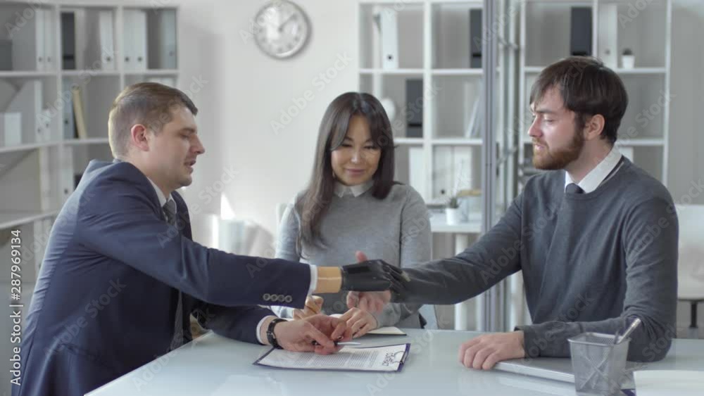 Caucasian entrepreneur with prosthetic hand talking and shaking hands with male business partner while their Asian female colleague writing in notepad during office meeting Caucasian entrepreneur with prosthetic hand talking and shaking hands with male business partner while their Asian female colleague writing in notepad during office meeting