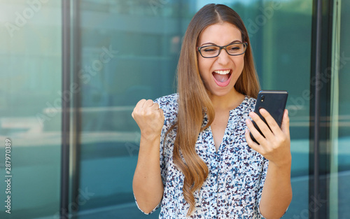 Excited young business woman receiving good news on smart phone celebrating with fist up outdoor. Copy space.