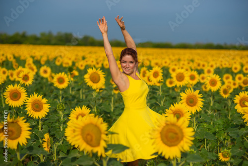 Red-haired woman in a yellow dress dancing with raised hands in a field of sunflowers. Beautiful girl in a skirt sun enjoys a cloudless day in the countryside. Pink locks of hair.