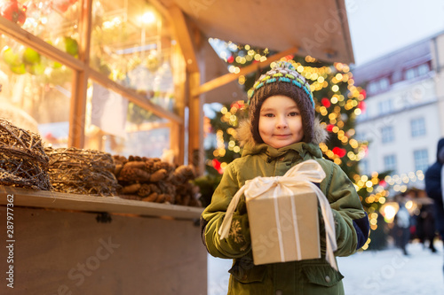 holidays, childhood and people concept - happy little boy with gift box at christmas market in winter evening